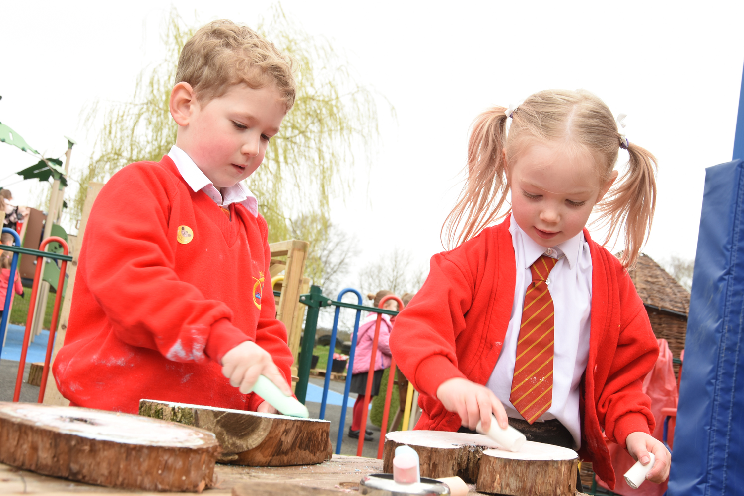 Two young students in red sweaters and ties engage in outdoor learning, using chalk to draw on tree stumps. The focus is on environmental education, plastic reduction, recycling, and litter picking.